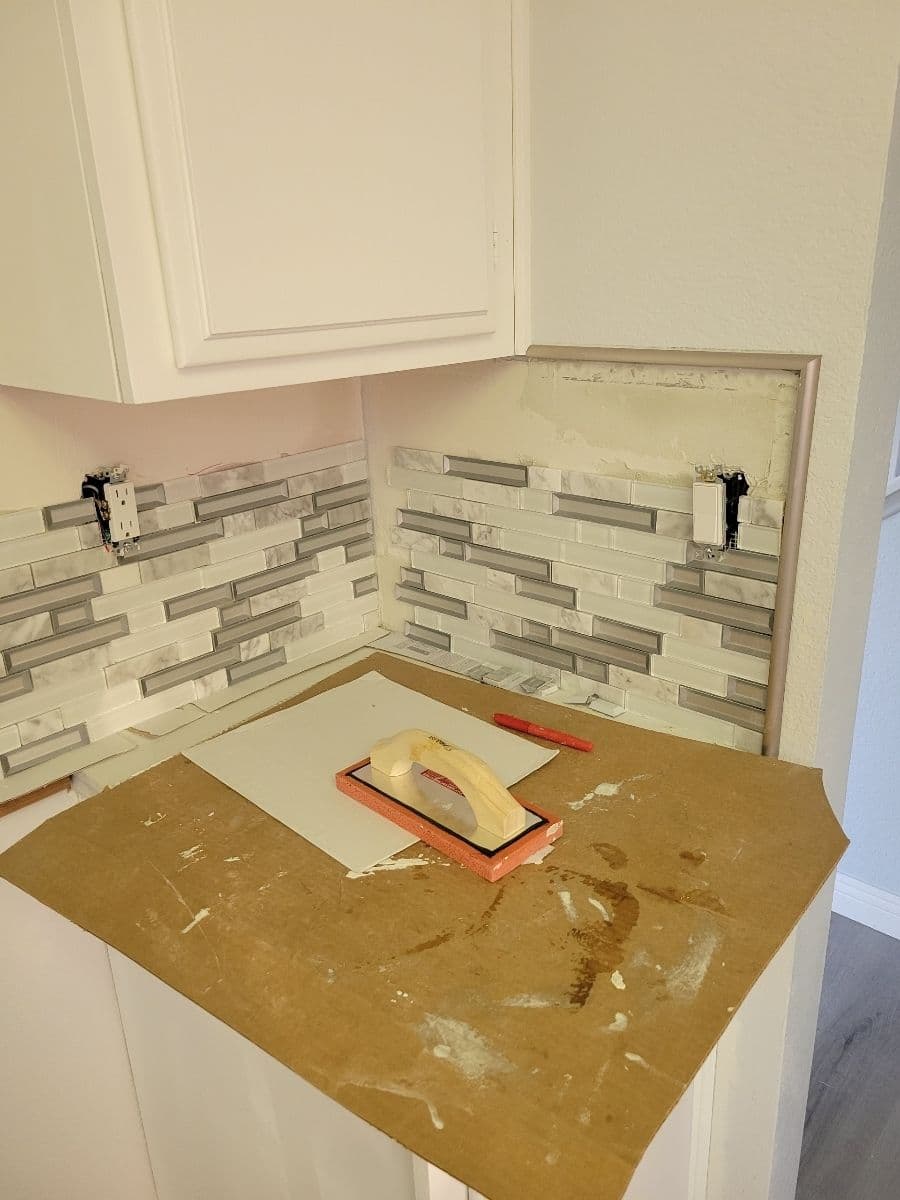 Kitchen backsplash installation with gray and white tile, tools, and materials on a counter.