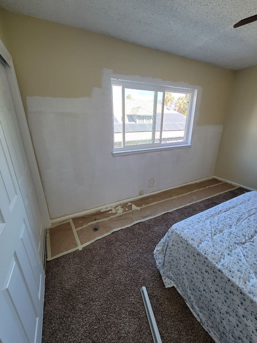 Bedroom with partially painted wall, carpeted floor, and a window overlooking rooftops.