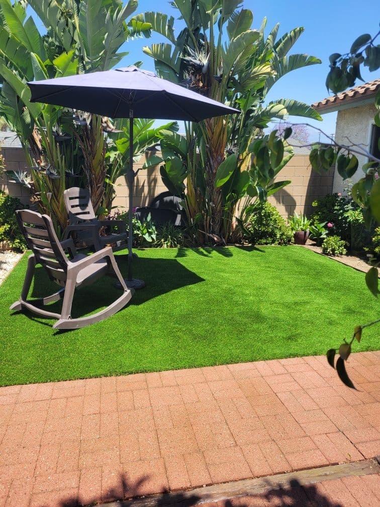 Relaxing backyard oasis with artificial grass, rocking chair, and sunshade surrounded by tropical plants.