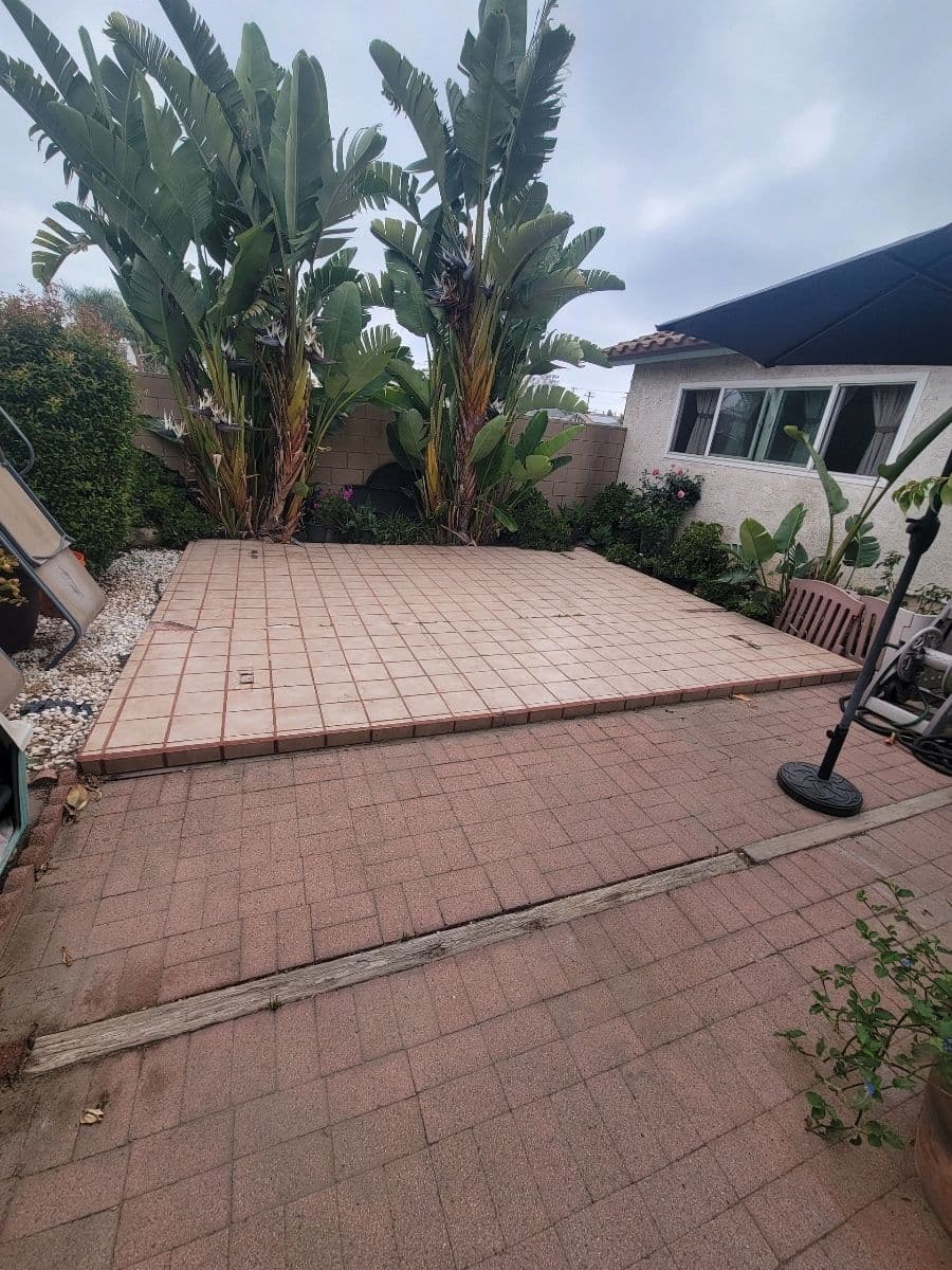 Paved outdoor patio surrounded by tropical plants and a house with a blue awning.